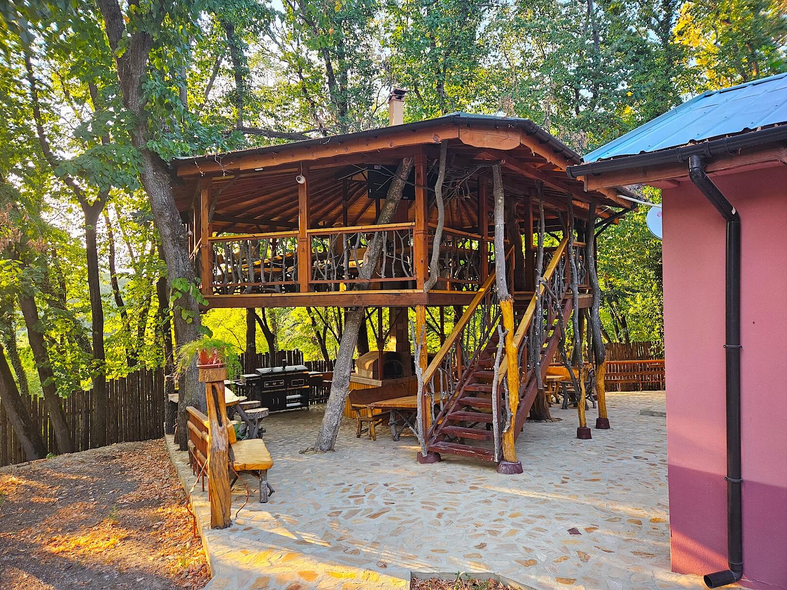 Alen Mak Hotel - A wooden treehouse with an open-air seating area is built around several trees, adjacent to a pink house. The surroundings are lush and green, reminiscent of cabins in the woods.